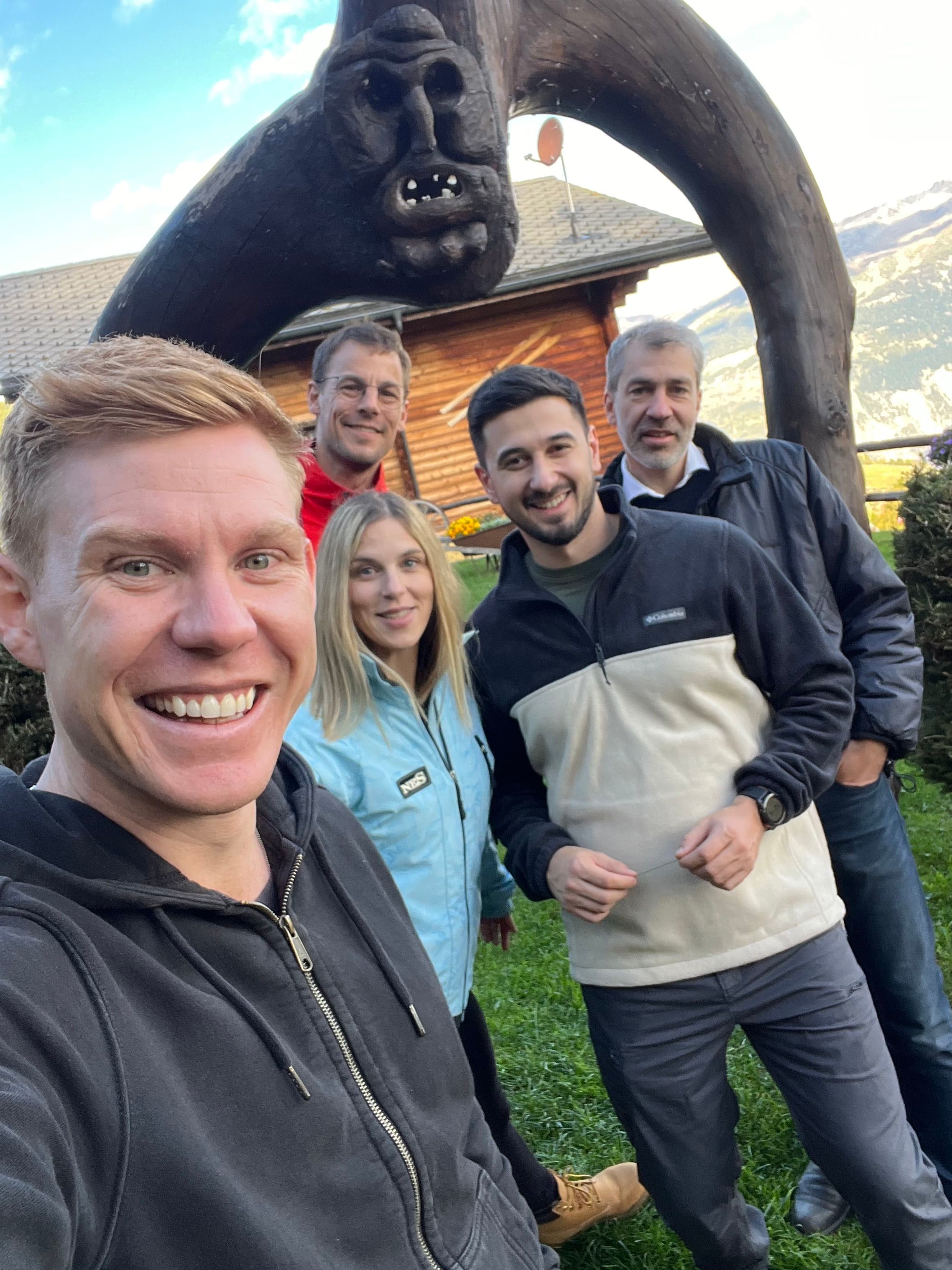 Five people smiling outdoors in front of a wooden sculpture with a cabin and mountains in the background.