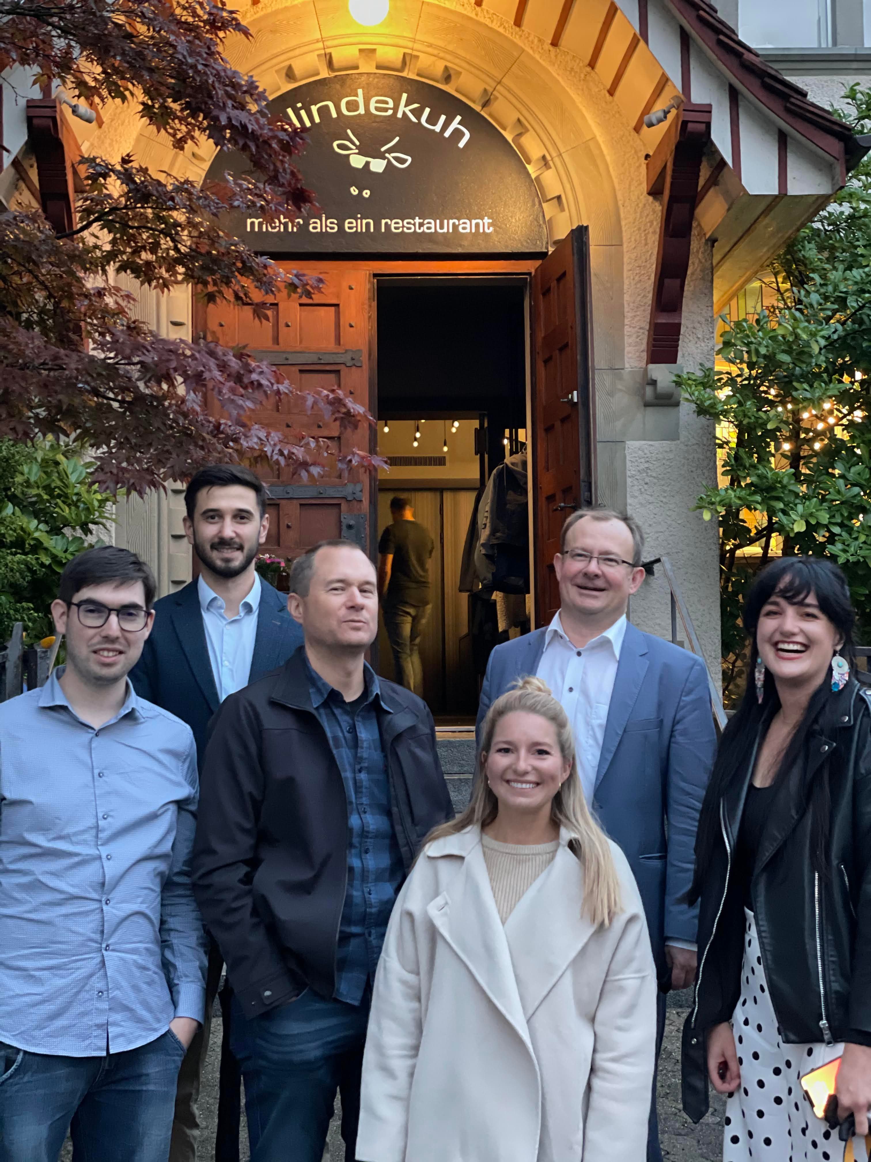 Group of seven people smiling in front of a restaurant entrance with a sign above.