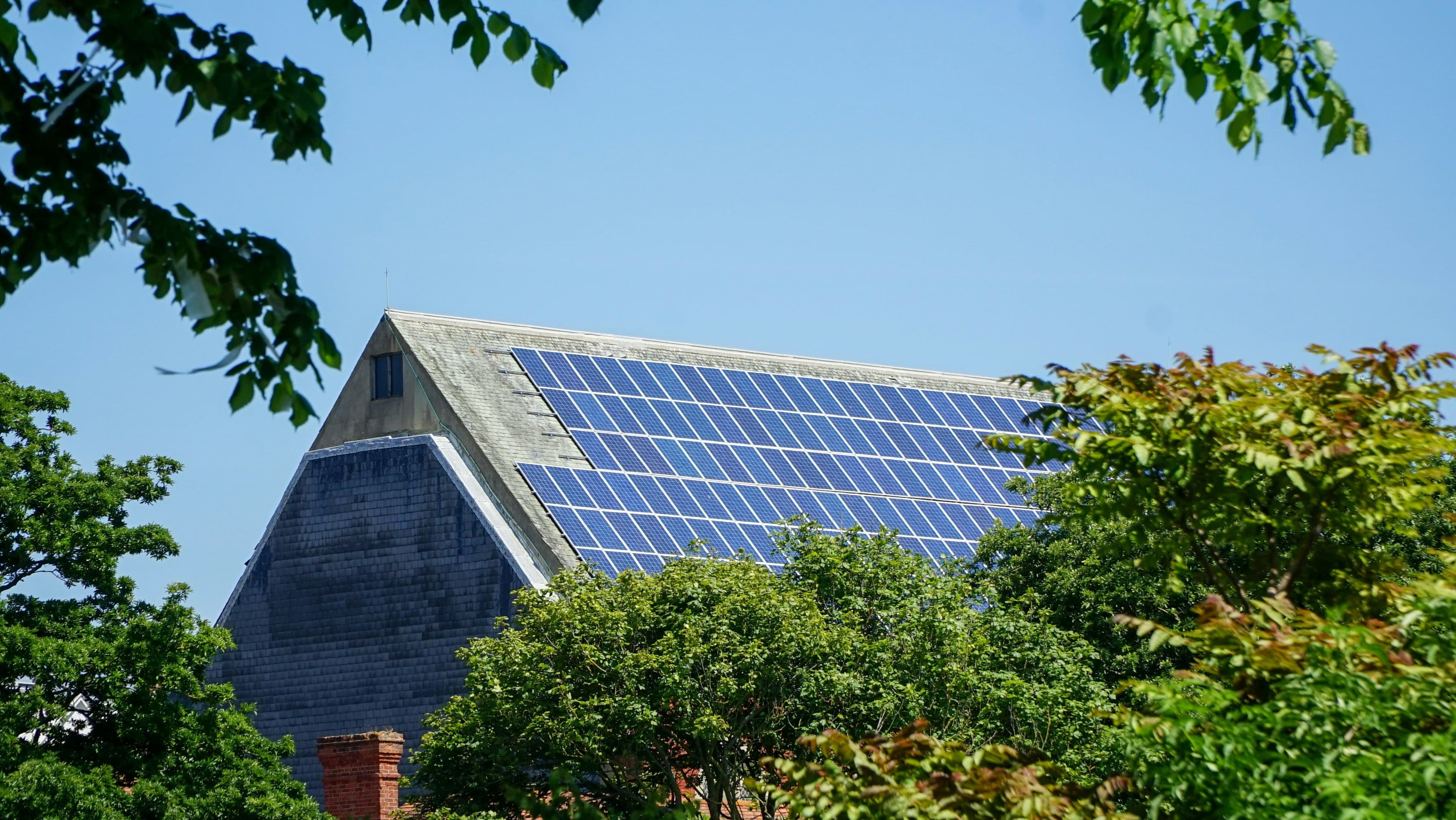 Solar panels on a large barn roof surrounded by lush green trees under a clear blue sky.