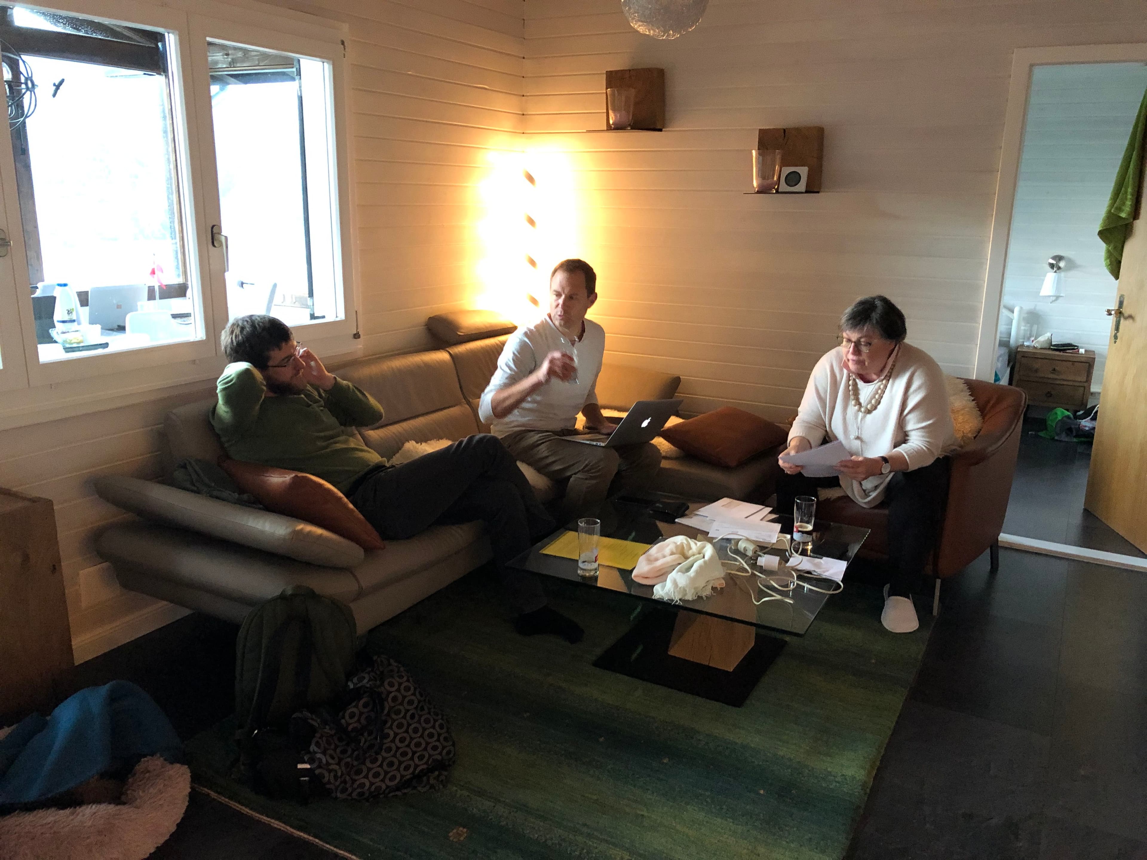 Three people in a cozy living room, sitting on a couch and chairs, engaged in conversation with papers and a laptop on a table.