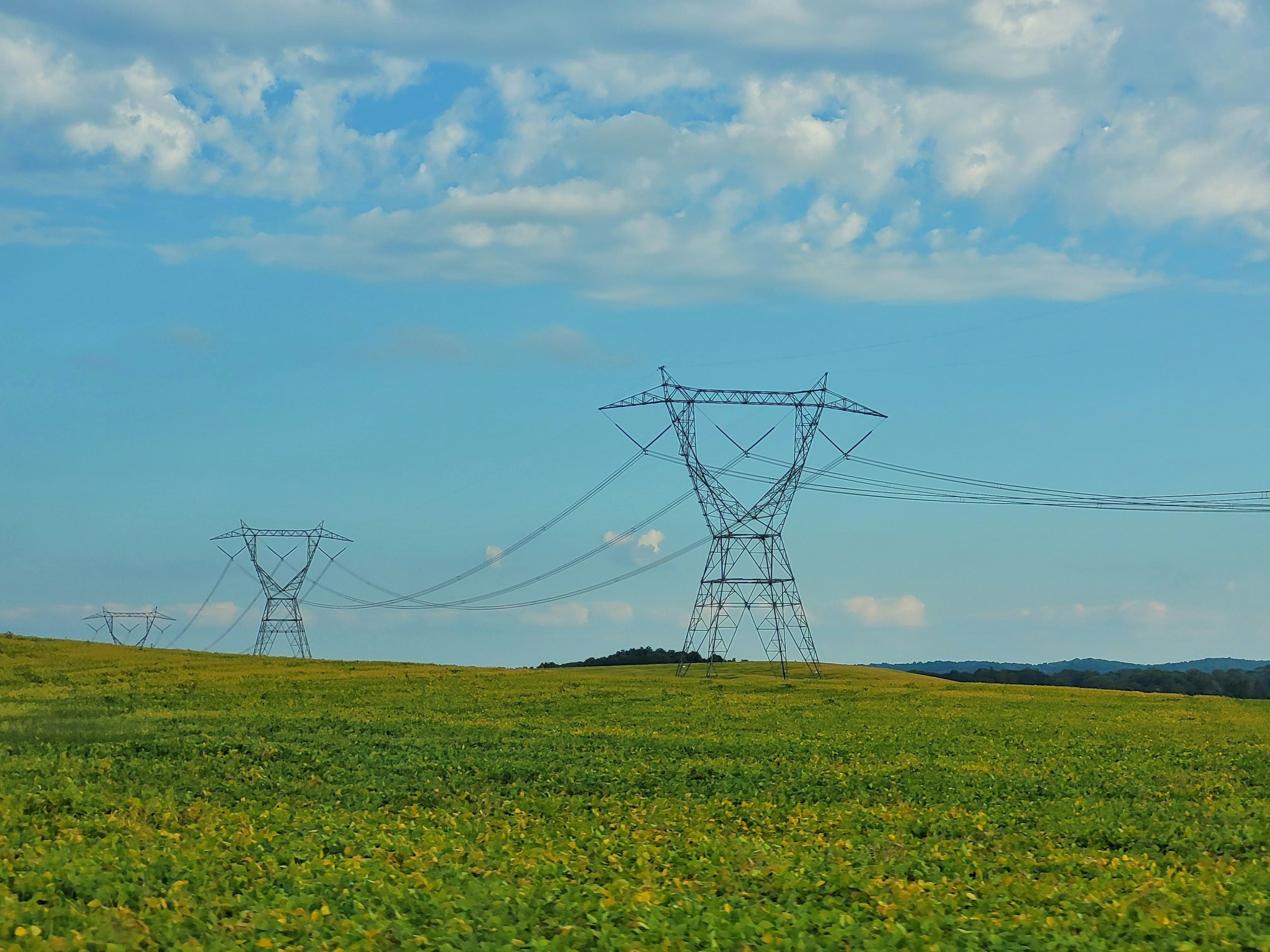 Power lines stretch across a green field under a blue sky with scattered clouds.
