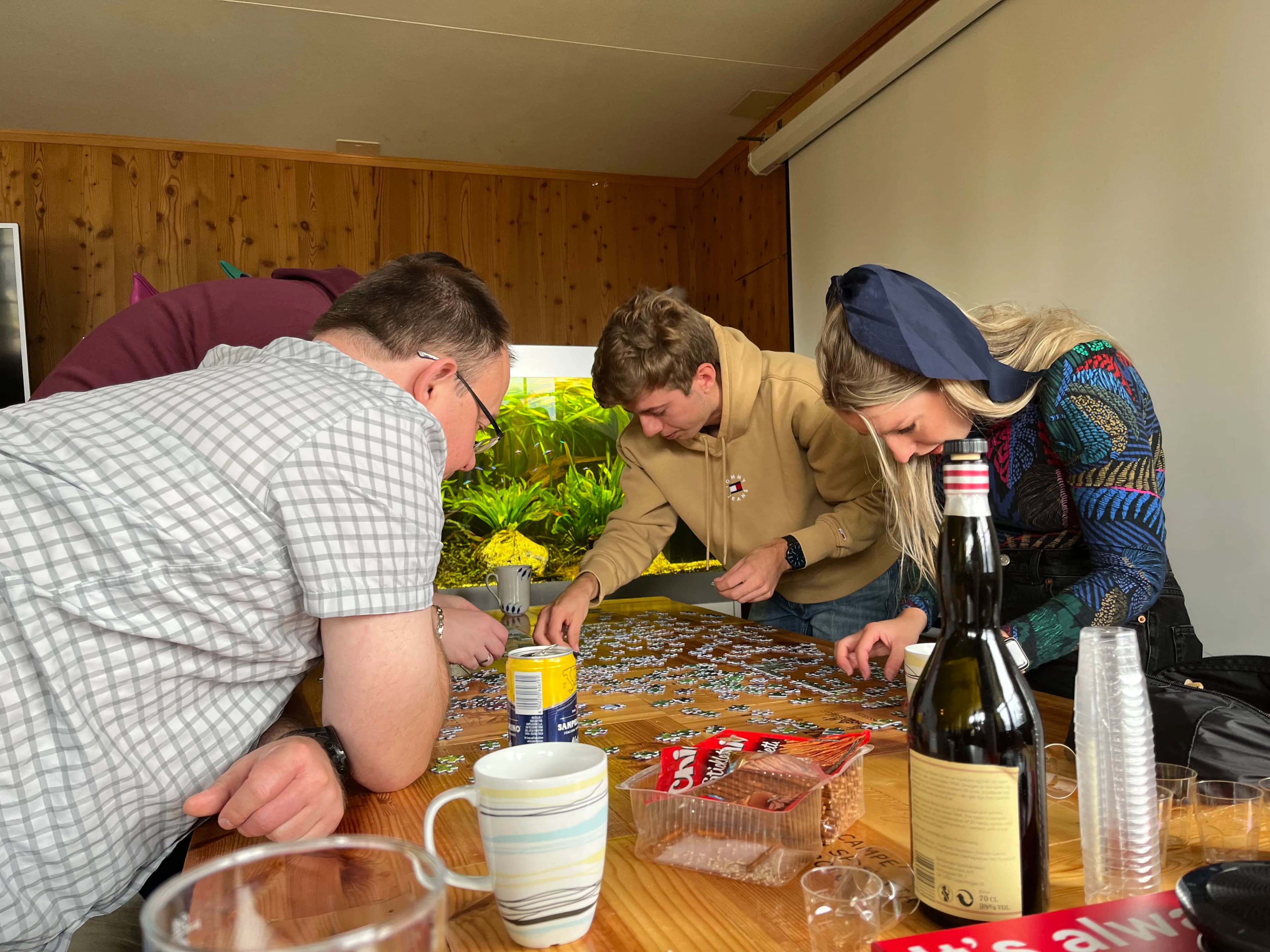 Four people are gathered around a table working on a jigsaw puzzle. Drinks and snacks are visible on the table.