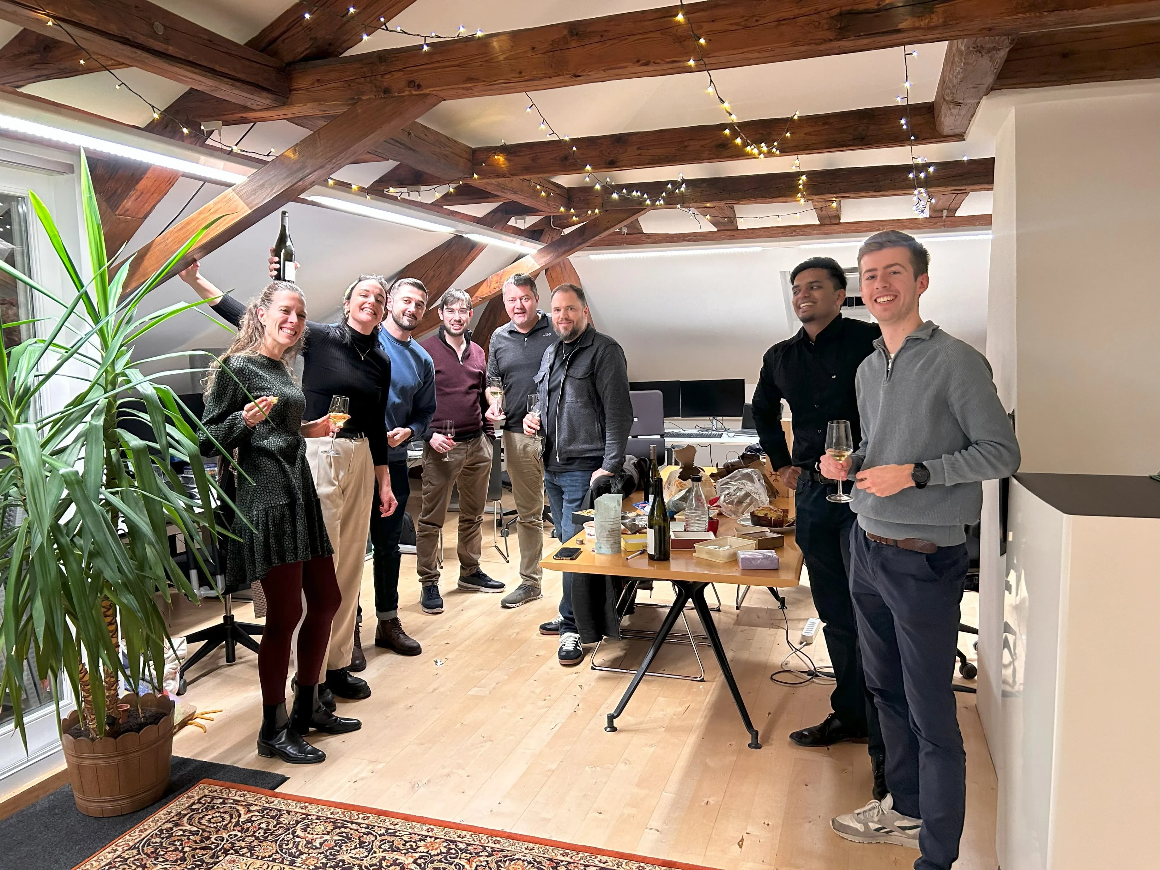 A group of people smiling and holding drinks in a cozy, decorated room with wooden beams and a large plant. A table with snacks is in the center.